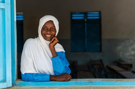 A young girl smiles at the camera, looking through an open window.