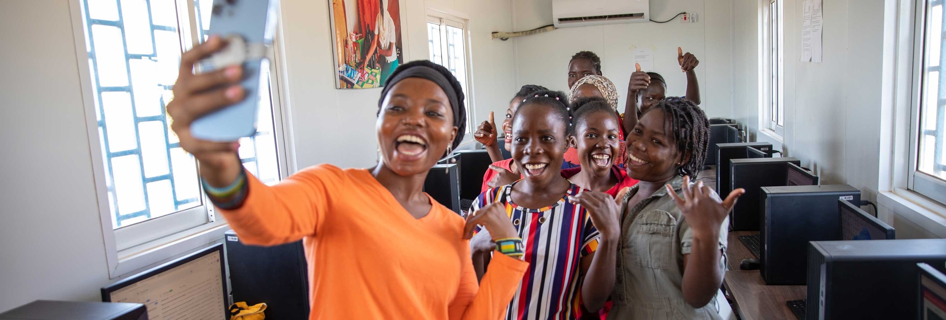 A woman in a bright orange shirt takes a selfie with a group of young women in a computer lab.