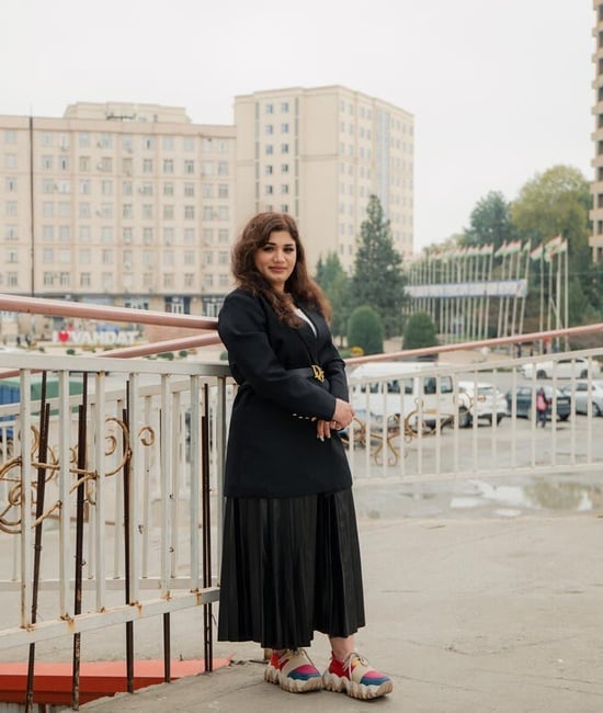 A young woman pictured leaning against a railing, with a carpark and tall buildings in the background