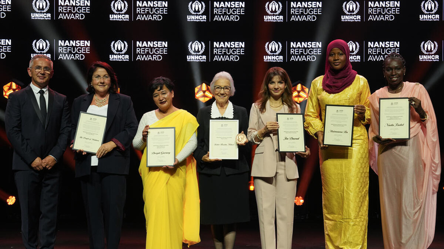 Seven people pose for a photo on a stage. They're holding framed certificates.