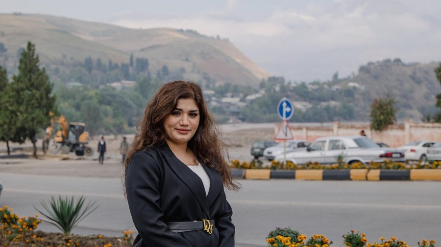 A smiling woman pictured in front of flowers alongside a road, with large hills in the distance.