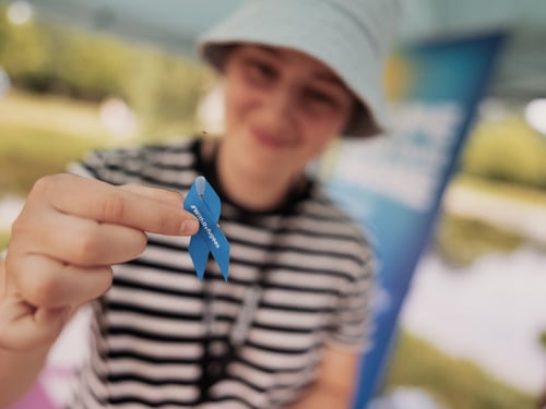 Une personne en tenue décontractée et portant un chapeau tient vers l’objectif un ruban bleu, tandis que son visage apparaît légèrement flou en arrière-plan