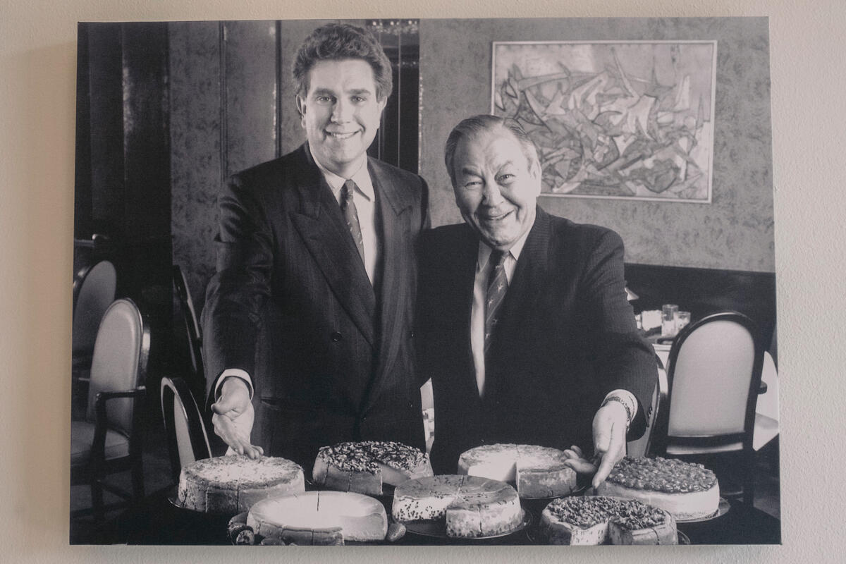 Photo en noir et blanc de deux hommes debout derrière une table chargée de différents gâteaux au fromage. 