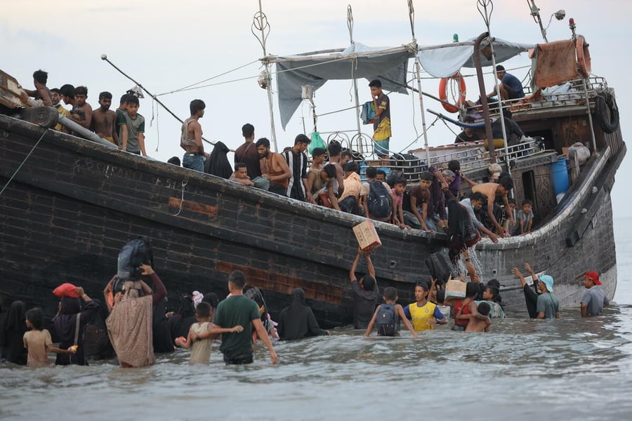 A crowded wooden boat is anchored in shallow coastal water as dozens of people disembark. Men, women, and children wade through the water carrying bags and boxes while others remain on deck, passing belongings down from the boat.