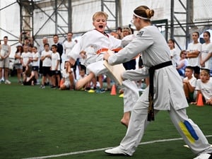 A 10 year old blond boy performing a taekwondo kick is in mid-air, preparing to break a wooden plank held by his instructor.