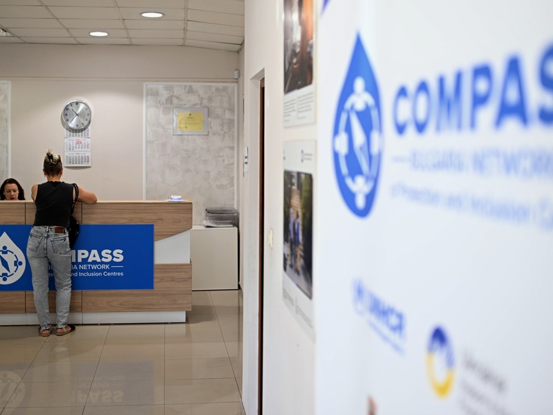 Photo shows the counter of a Compass centre, we see one woman with her back turned to the camera talking to the woman receptionist. A large sign "COMPASS" is visible on the right.
