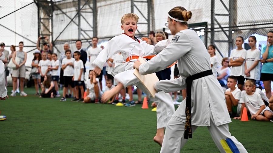 A 10 year old blond boy performing a taekwondo kick is in mid-air, preparing to break a wooden plank held by his instructor.