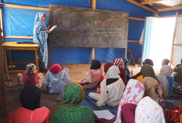 A woman is standing next to a chalkboard, in a classroom full of young girls.
