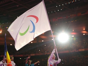 A woman wearing glasses holds a large IPC flag above her head in a brightly lit stadium.