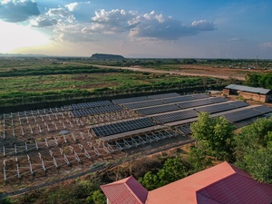 An aerial view of a partially constructed solar farm with a red-roofed building in the foreground and a savannah landscape stretching to the horizon.  