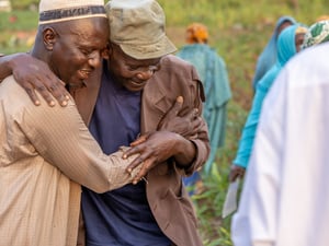 Two men smile and embrace in a field, surrounded by other people