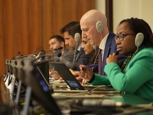 Close up of five speakers in a conference room, pictured with headphones on and laptops and microphones in front of them