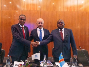 Two men in suits shake hands across a third man behind a desk