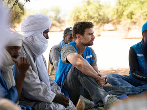 A man wearing a blue UNHCR vest sits on the ground outside next to men wearing white headscarves.