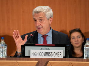 Un homme est assis derrière une estrade et prononce un discours. Une pancarte portant l'inscription « High Commissioner » (Haut Commissaire) est placée devant lui.