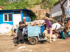 A man and several children sit and stand near their belongings outside in front of a shelter.