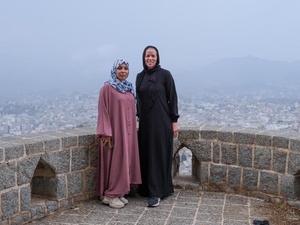 Two women stand on a stone terrace with a city landscape behind them