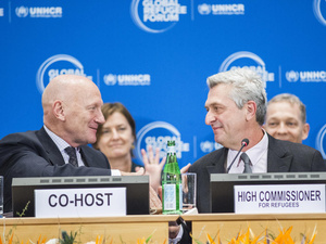 Two men shake hands on a podium behind the signs 'Co-host' and 'High Commissioner for refugees'.