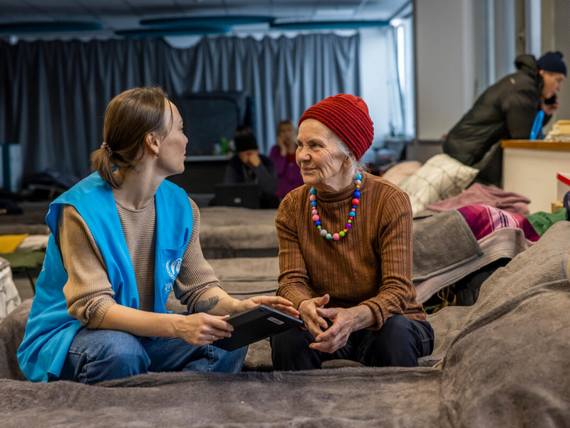 UNHCR staff member discussing with an older woman, both sitting on a bed.