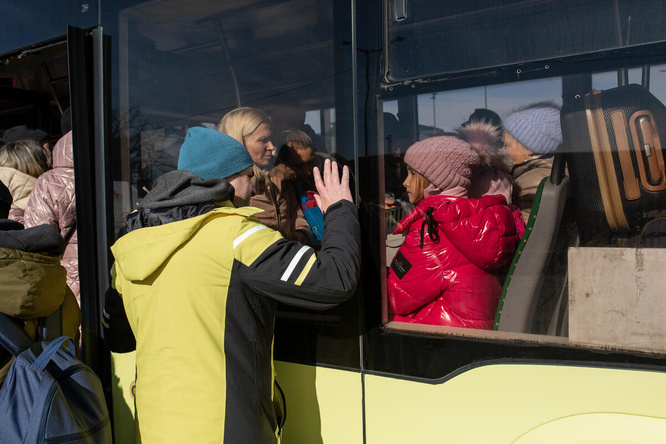 A man waves to a young girl sitting on a bus, they are separated by the window glass.