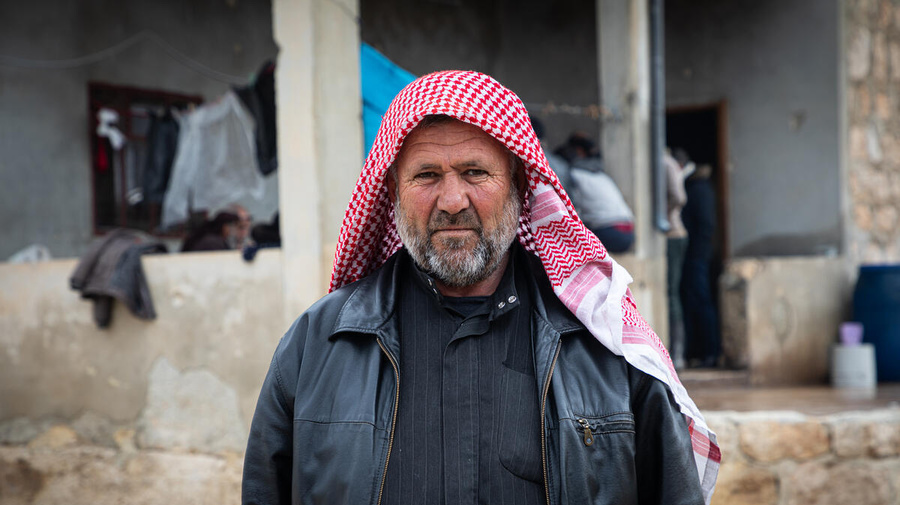 A man wearing a red headscarf and leather jacket stands in front of a building.