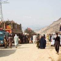 Afghan returnees walk along a dirt road beside loaded trucks.