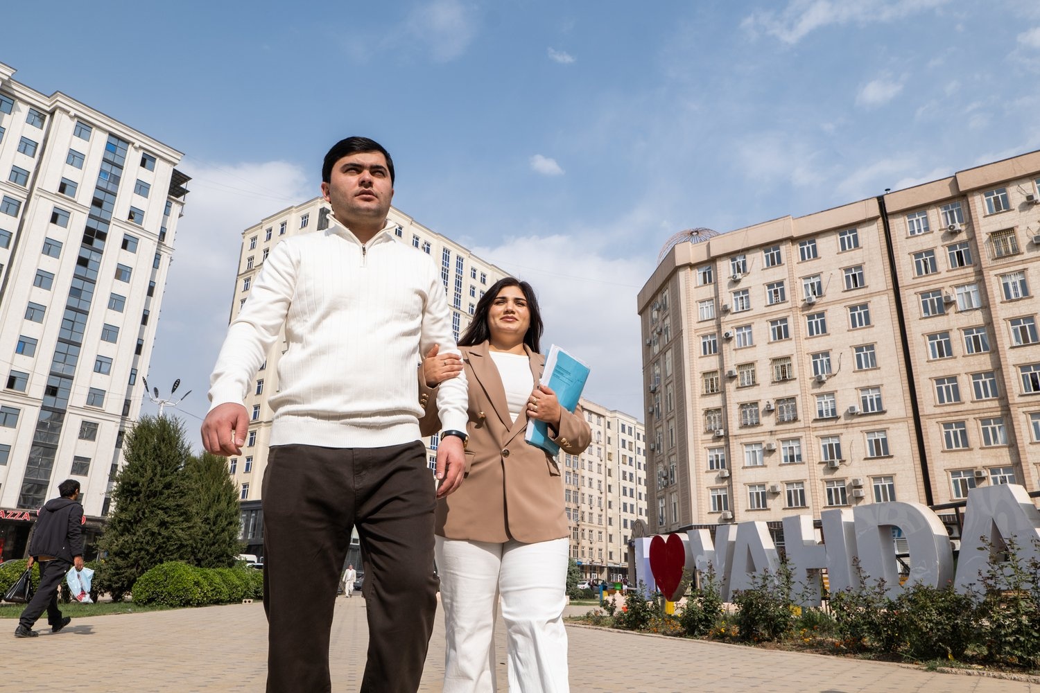 A couple walks arm in arm in a square backed by tall apartment buildings