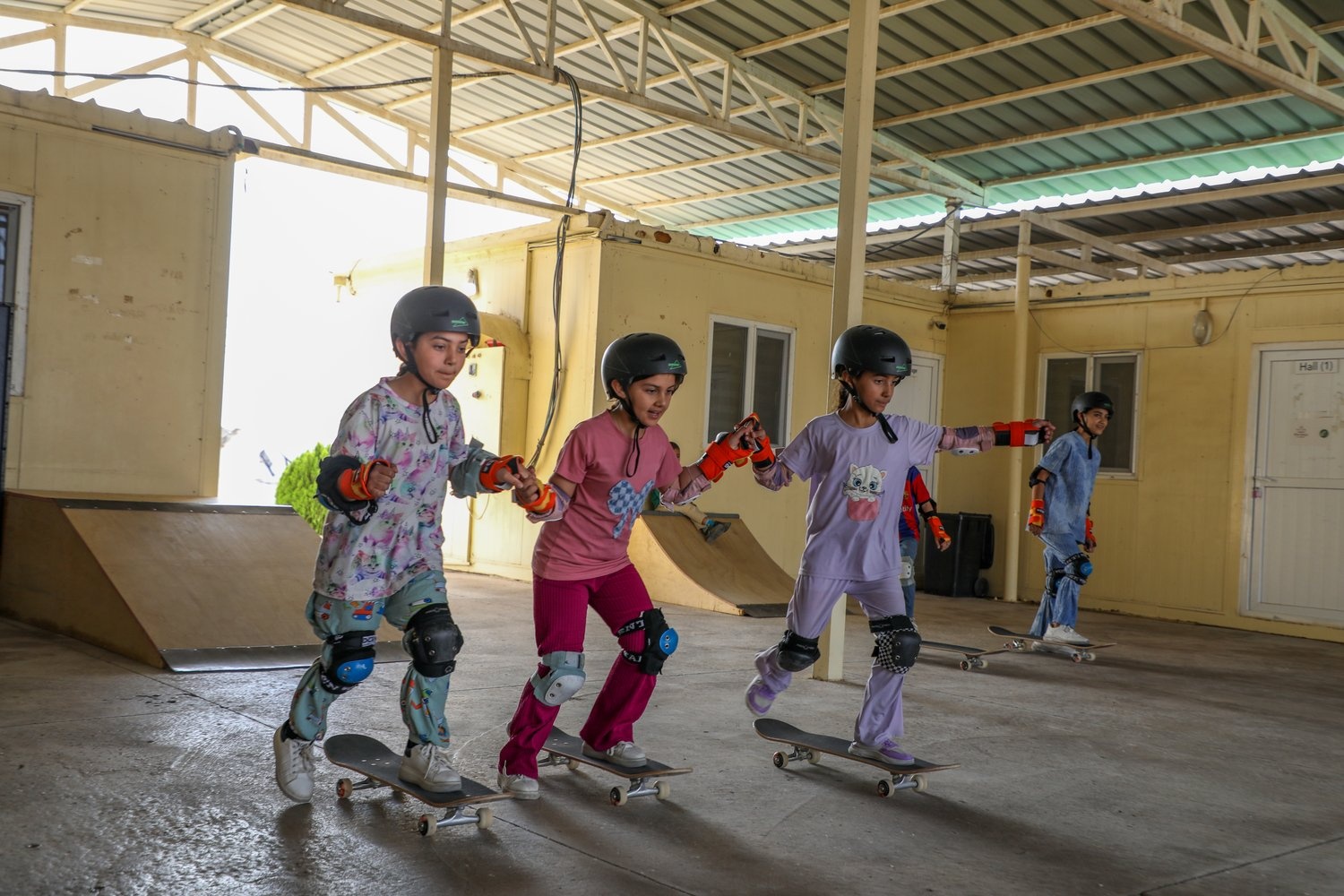 Girls wearing protective equipment ride skateboards inside a large building