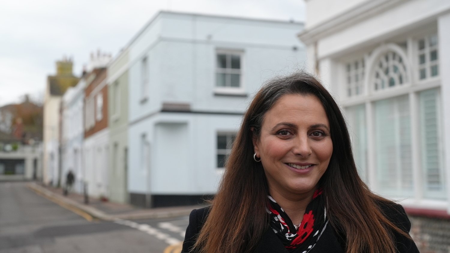 A woman in a black jacket and bright scarf stands in a residential street 
