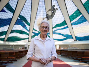 A woman with white hair weaing glasses and a white shirt stands inside a large glass-domed church.