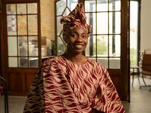 A smiling woman in a patterned traditional dress and headscarft sits in an office