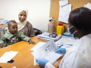 A woman is seated at a table with her young son, across from a medical staff member staff in a mask who is looking at paperwork