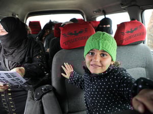 A young girl wearing a knitted green hat sits inside a minibus with women passengers sitting in the seats behind her.