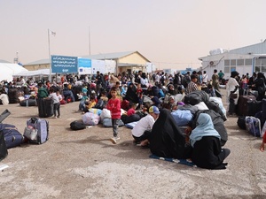 Families with their belongings gather at a border crossing, standing or sitting on the ground.