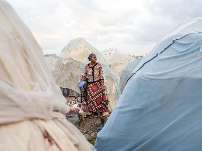 A woman stands in front of a makeshift shelter, surrounded by similar shelters.
