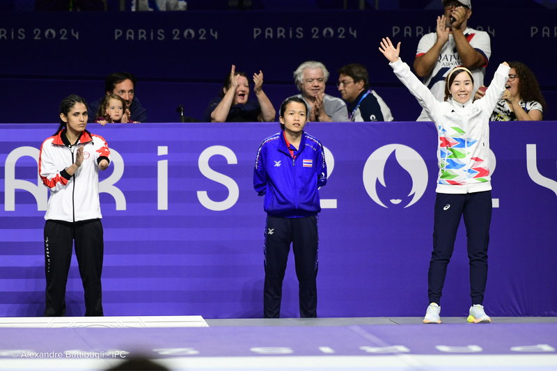 Une athlète lève les bras en signe de célébration, tandis que deux autres personnes applaudissent en regardant.