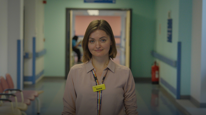 A woman stands in a hospital corridor wearing a yellow name tag reading 'Inna'