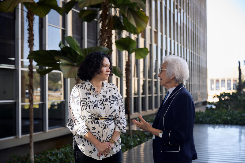 Two women talk on a walkway bordered by plants outside a large institutional building