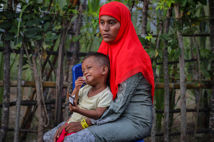 A Rohingya refugee woman holding her young son on her lap, looking into the distance.