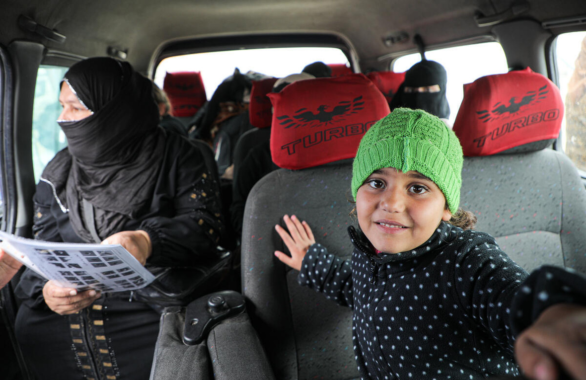 A young girl wearing a knitted green hat sits inside a minibus with women passengers sitting in the seats behind her.