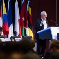 A man in a suit gestures while speaking at a lectern in front of a crowd