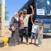 A couple and their three children stand on the roadside, smiling and waving in front of a bus with a UNHCR and IOM sign in the window. 