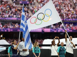 A man carries the Olympic flag in a packed stadium alongside a woman holding a Greek flag and others