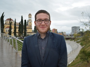 Alexander stands on a pedestrian bridge over the Gjanica River in Fier, Albania. Behind him is the Saint George Orthodox Cathedral. He smiles gently, relaxed, quietly confident, and fully present.