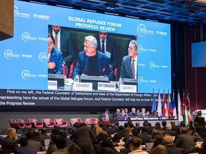 A man is pictured speaking on a giant screen in front of a conference room audience