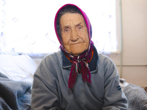 Portrait of older woman sitting on a bed, wearing scarf tied under her chin.
