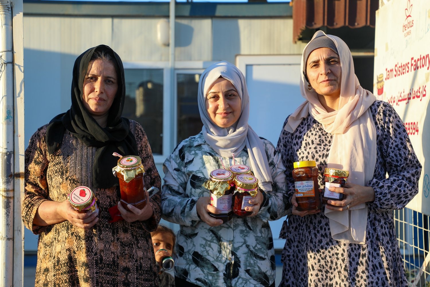 Three women stand outside a temporary building in the evening sunlight holding jars of homemade jam 