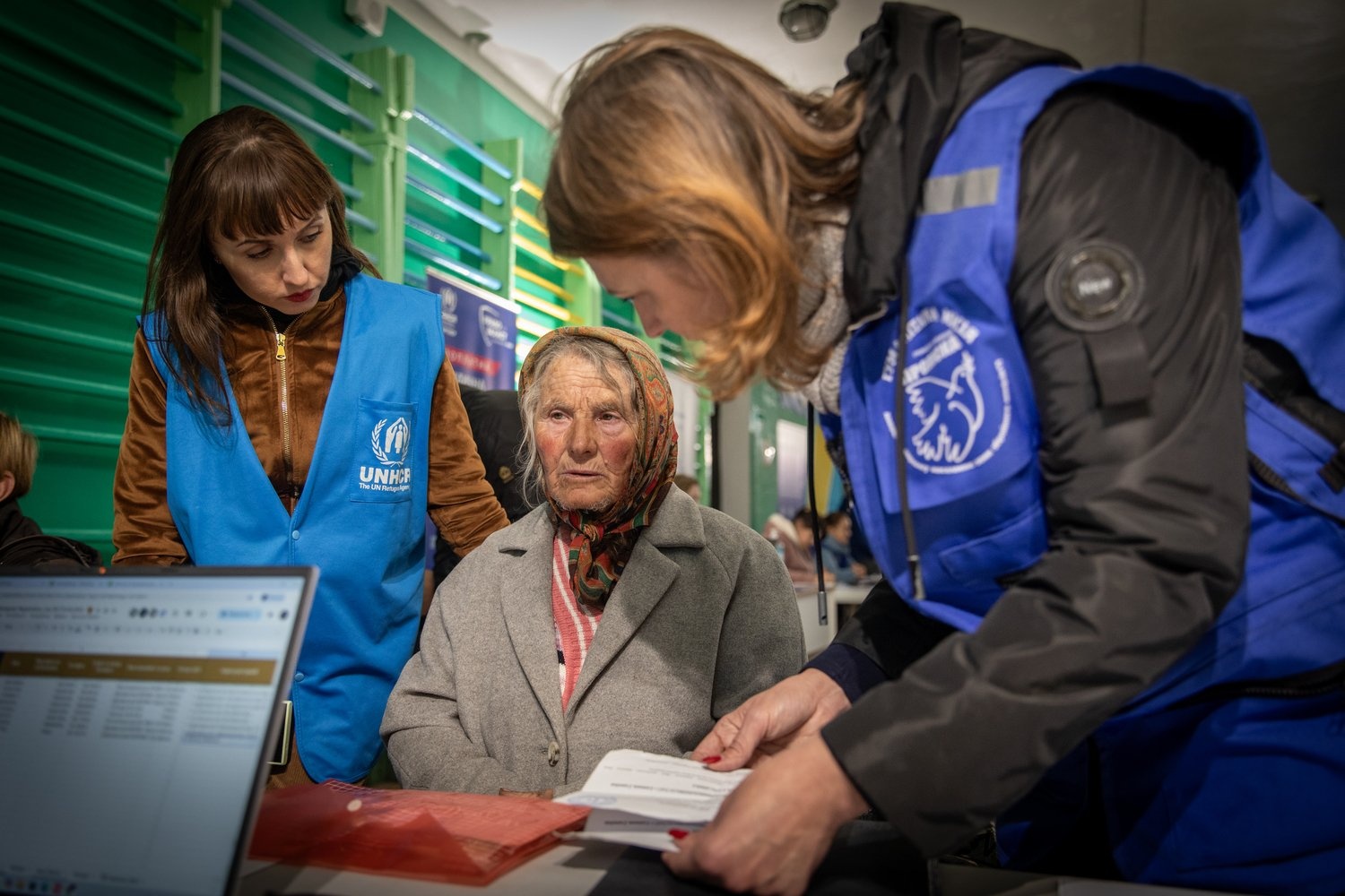 Two aid workers stand beside an elderly woman seated in front of a desk