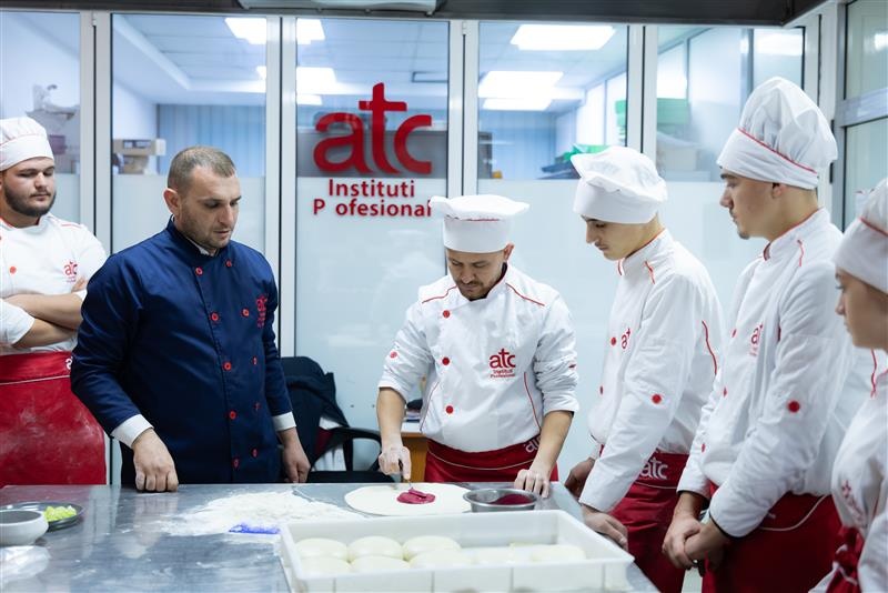 A man in a blue chef uniform instructs trainees in white chef uniforms.