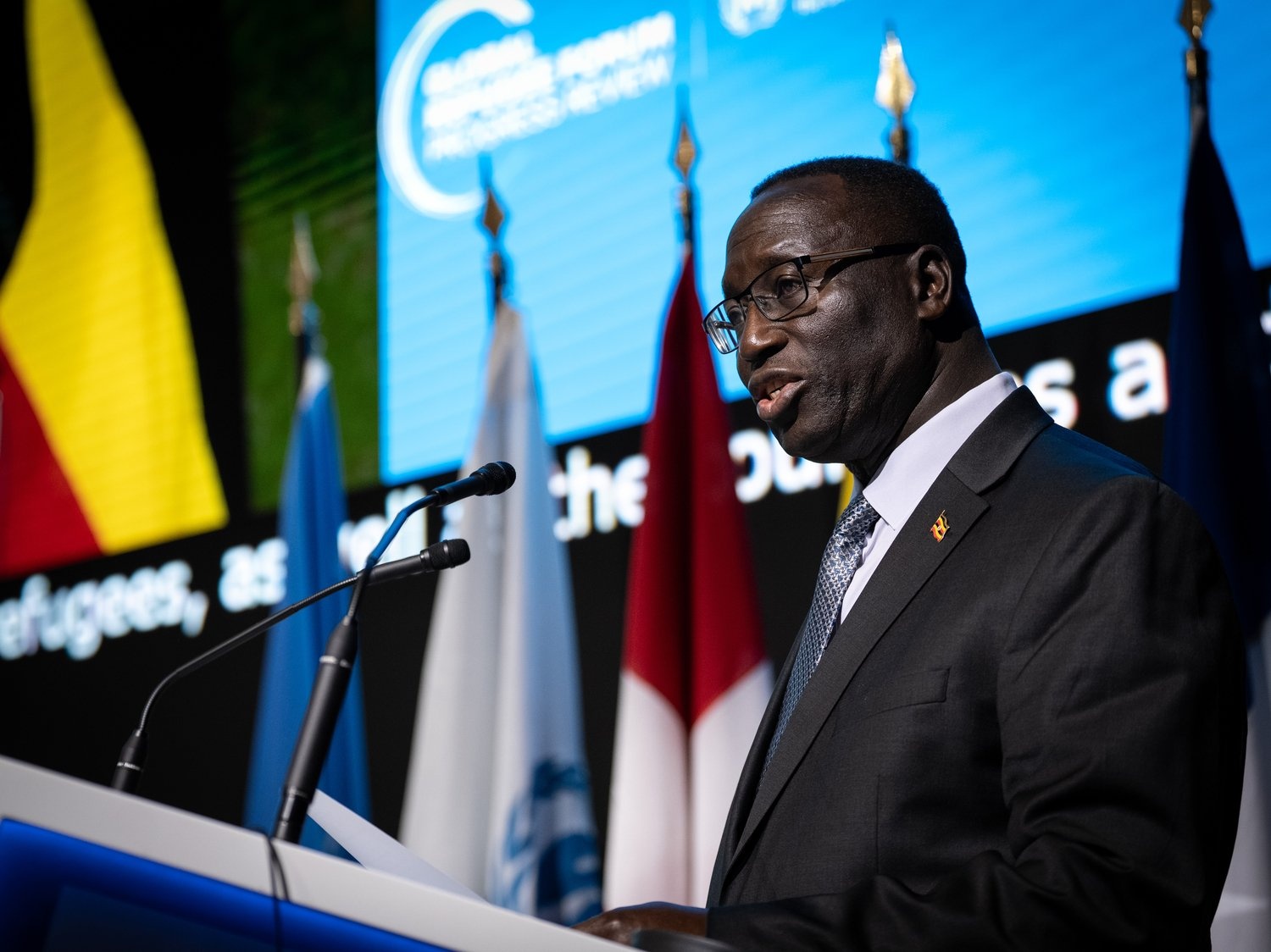 A man in a suit speaks at a lectern with flags and a giant screen in the background